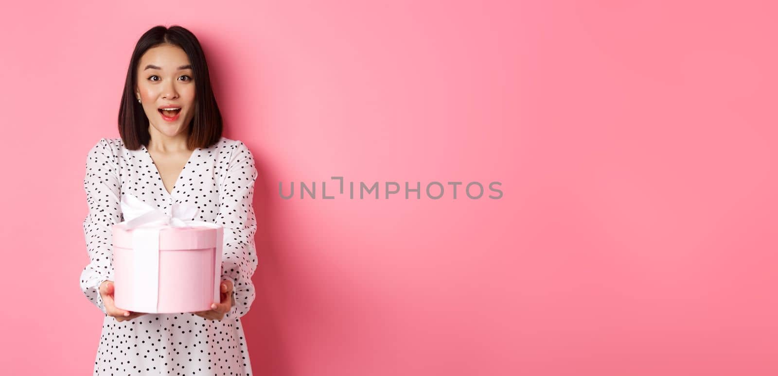 Cute asian woman congratulate with holiday or birthday, giving gift in cute box, standing over pink background.