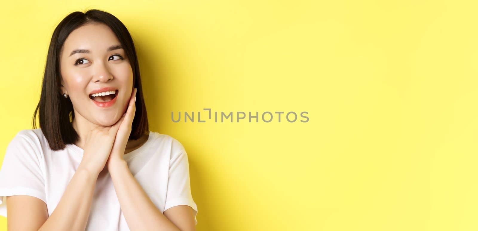 Cute romantic girl in white t-shirt imaging valentines day, looking left with admiration and joy, smiling with hands pressed to cheek, standing over yellow background.