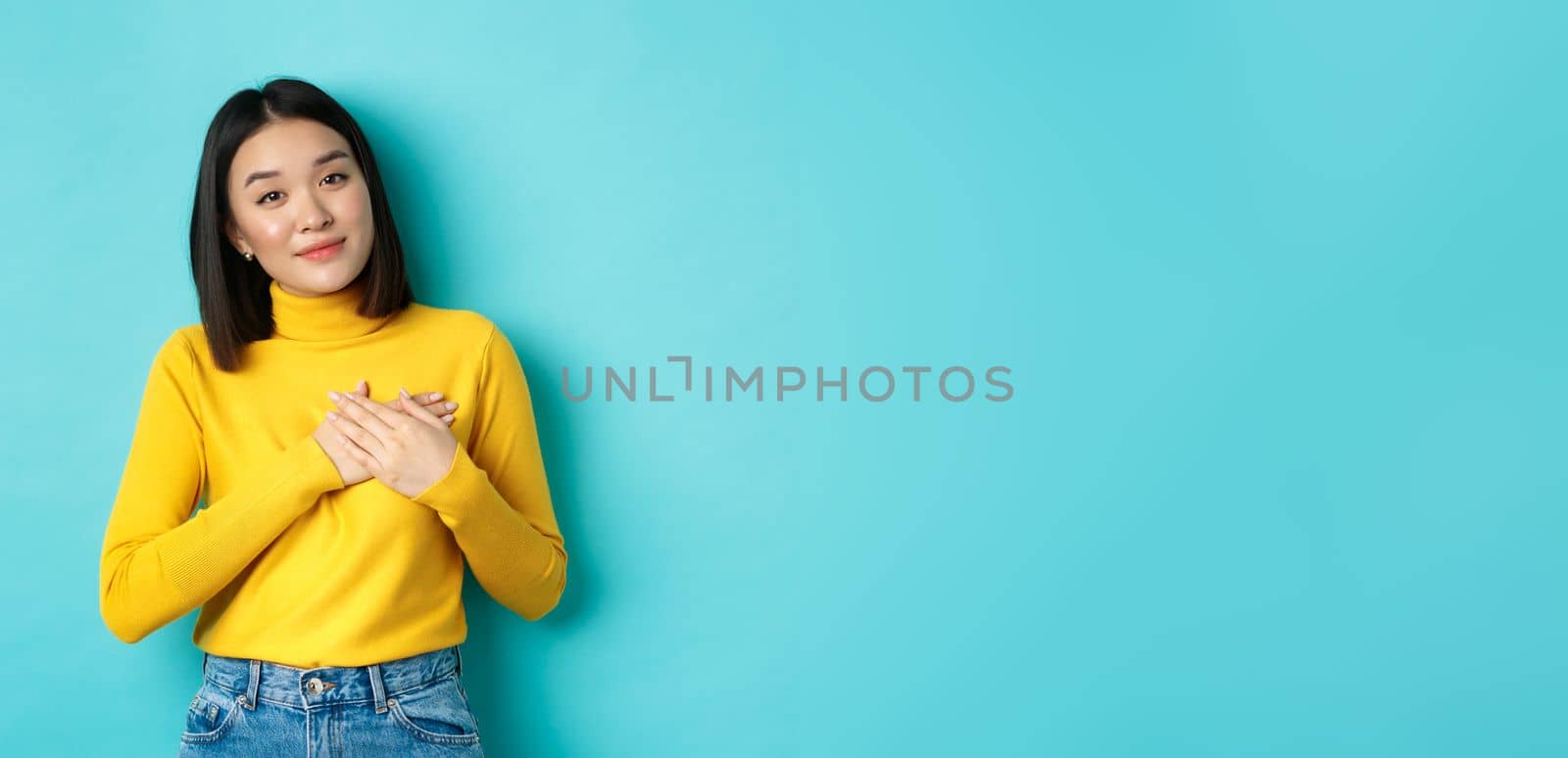 Portrait of beautiful heartfelt woman holding hands on heart, smiling and listening compassionate, standing over blue background in yellow pullover.