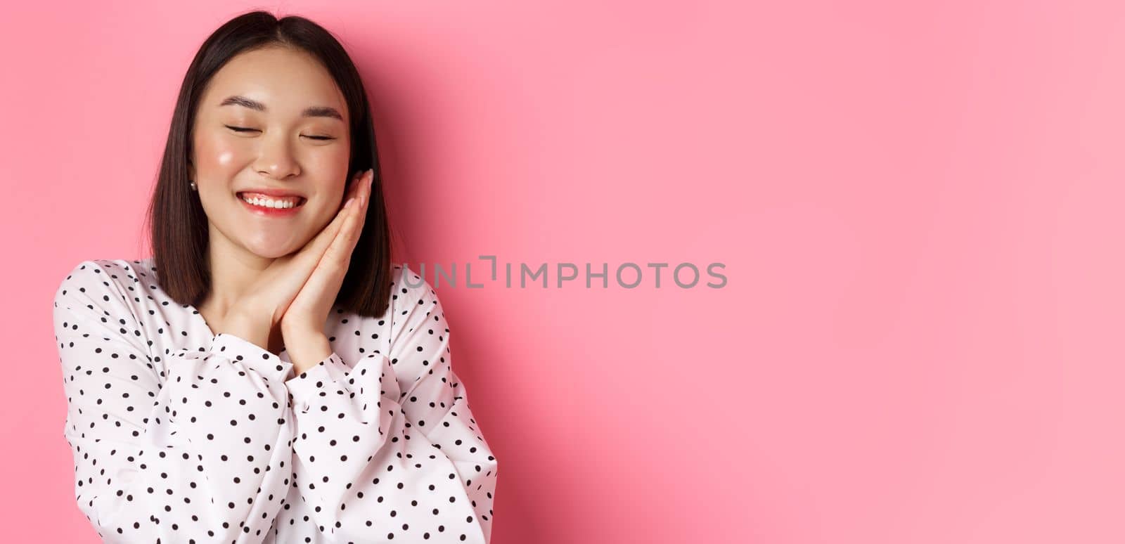 Beauty and lifestyle concept. Close-up of beautiful and dreamy asian woman sleeping on her hands, close eyes and smiling, daydreaming on pink background by Benzoix