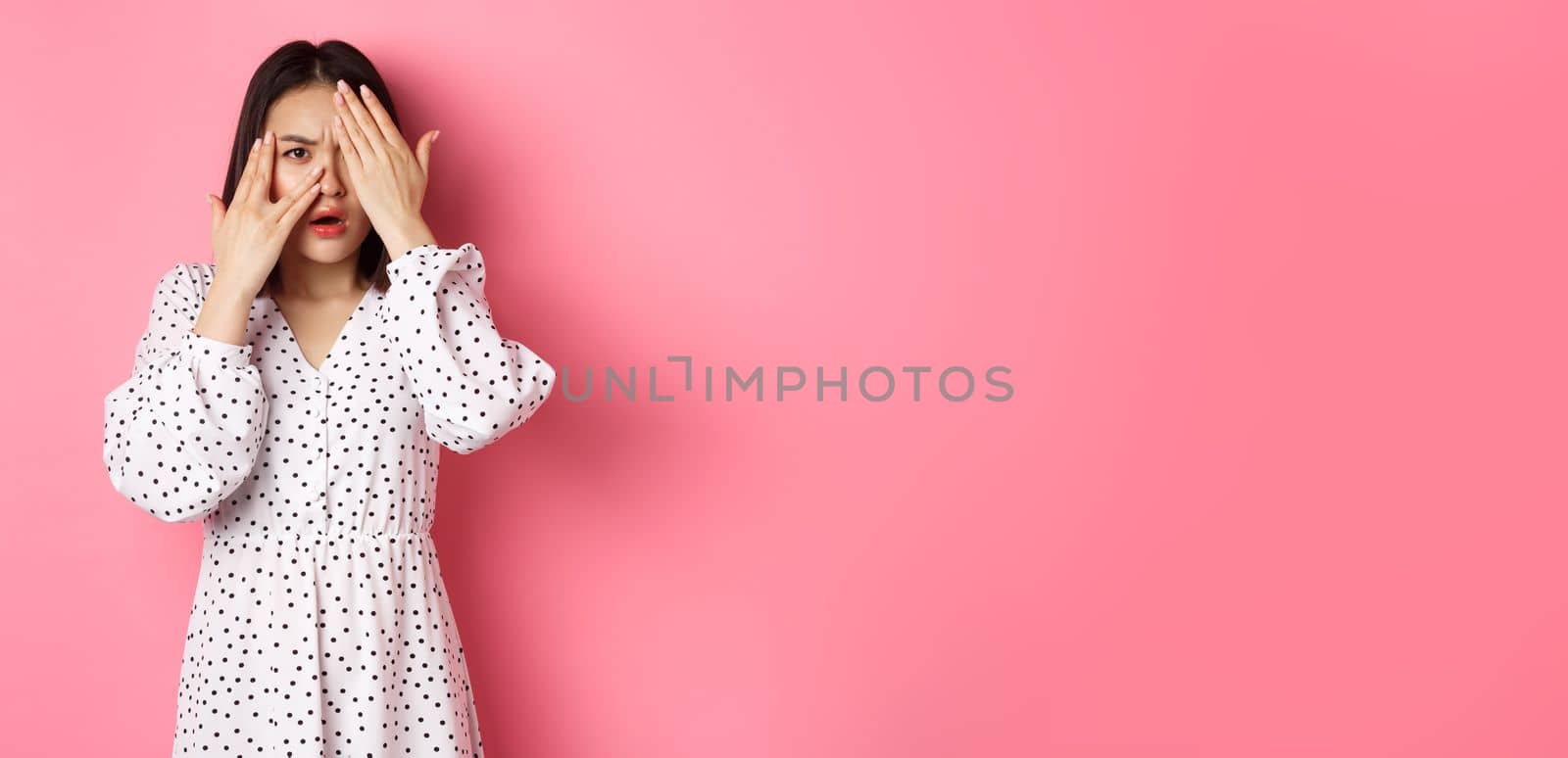 Disappointed asian woman peeking through fingers, open eyes and frowning displeased, staring with disdain at camera, standing over pink background by Benzoix