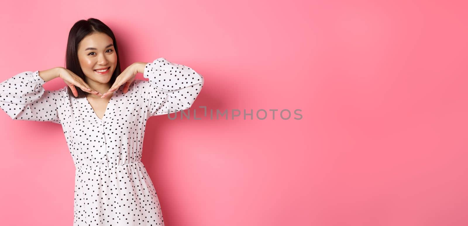 Lovely asian woman showing her beautiful clean face, smiling pretty, standing in dress against pink romantic background.
