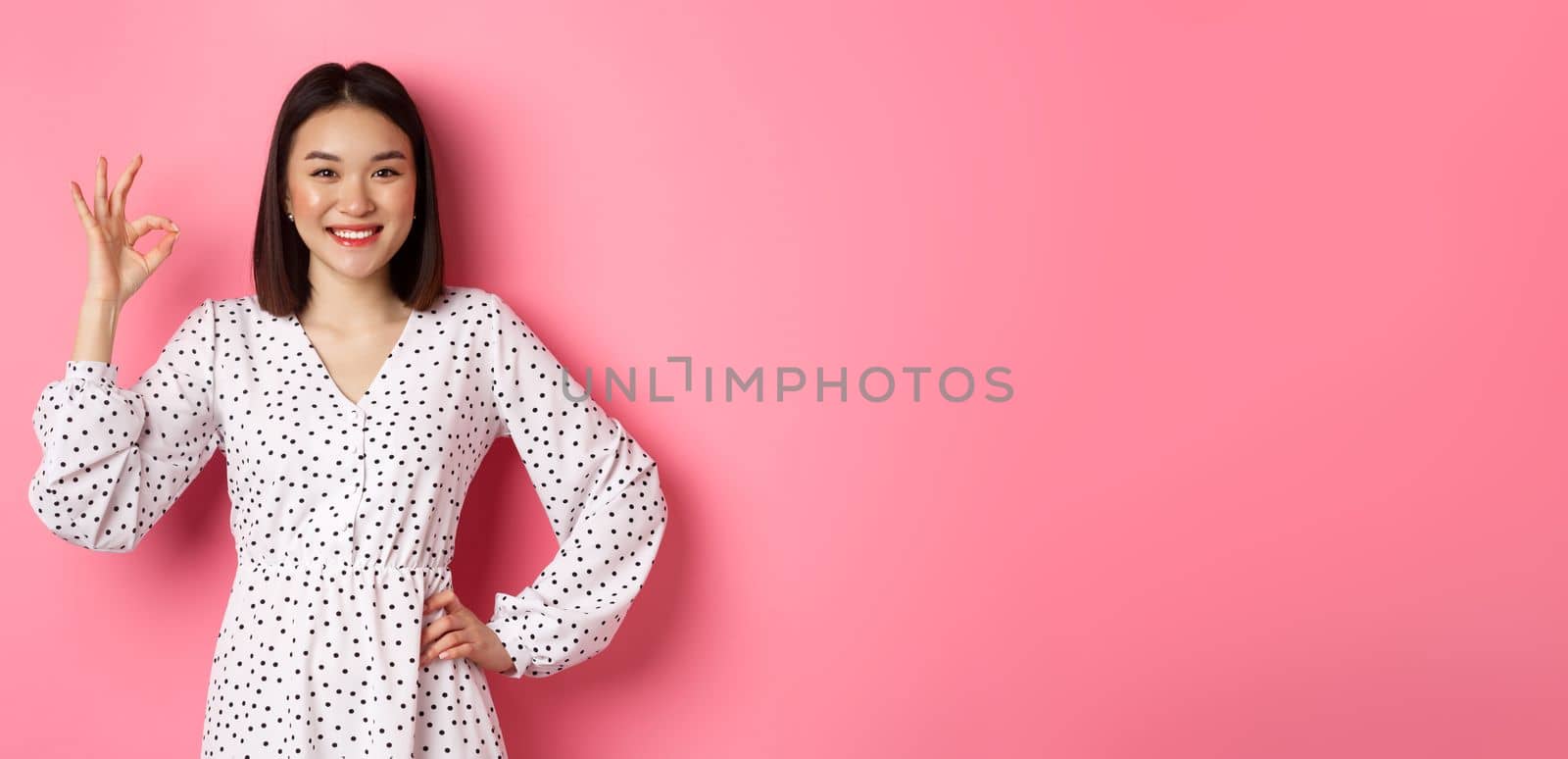 Pretty young asian woman in dress showing okay sign, praising and showing approval, looking satisfied, standing against pink background.