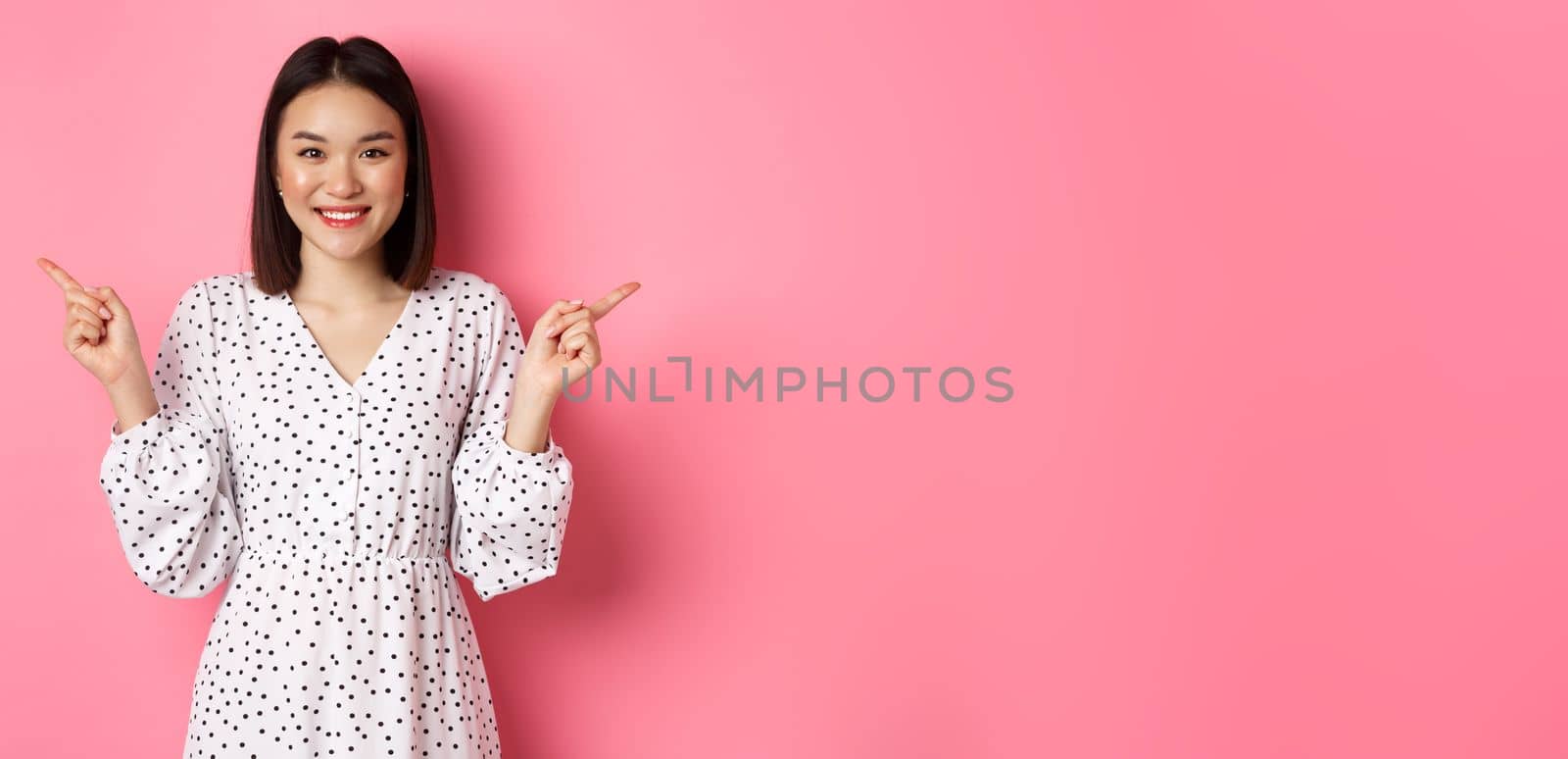 Beautiful asian woman making choice on shopping, pointing fingers sideways and showing variants, smiling at camera, standing over pink background by Benzoix