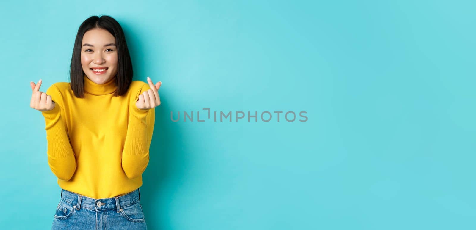 Beautiful asian girl in yellow sweater, smiling and showing finger hearts, standing happy against blue background.