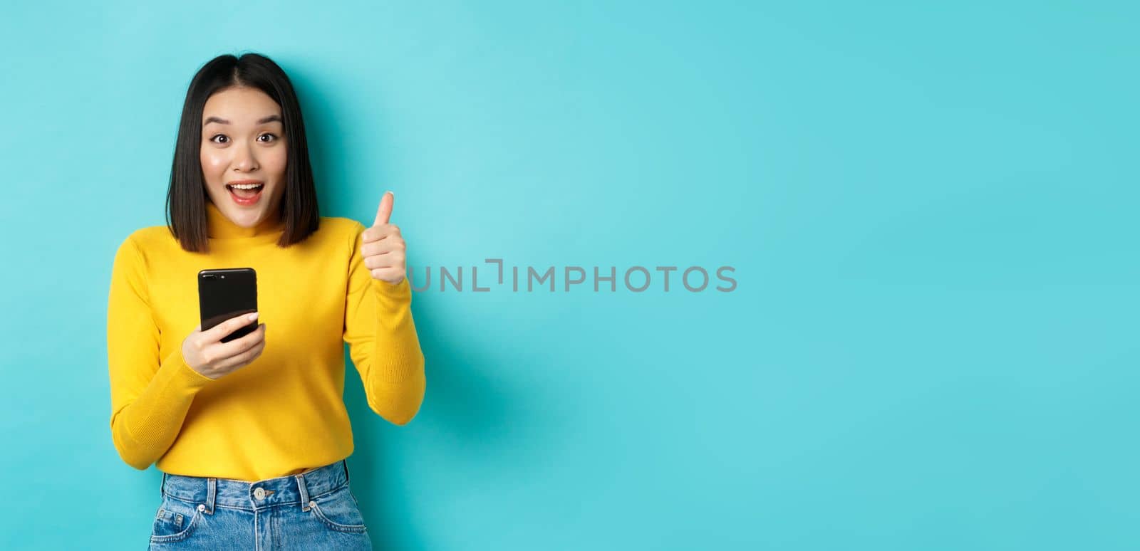 E-commerce and online shopping concept. Excited and amazed asian woman showing thumbs up after using smartphone app, recommend device, standing over blue background by Benzoix