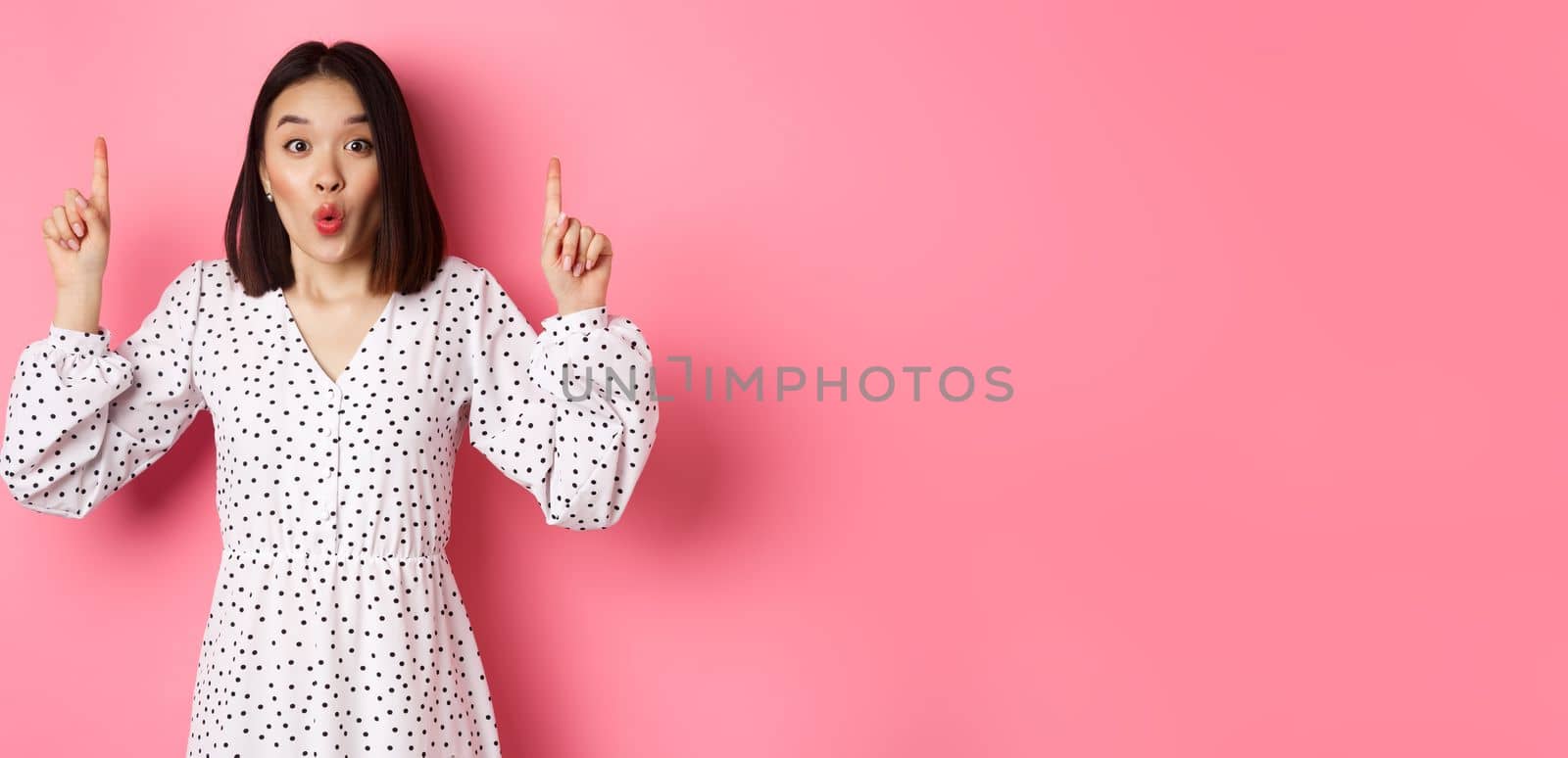 Pretty asian female model in dress pointing fingers up, showing copy space and staring at camera amazed, telling big news, standing over pink background by Benzoix