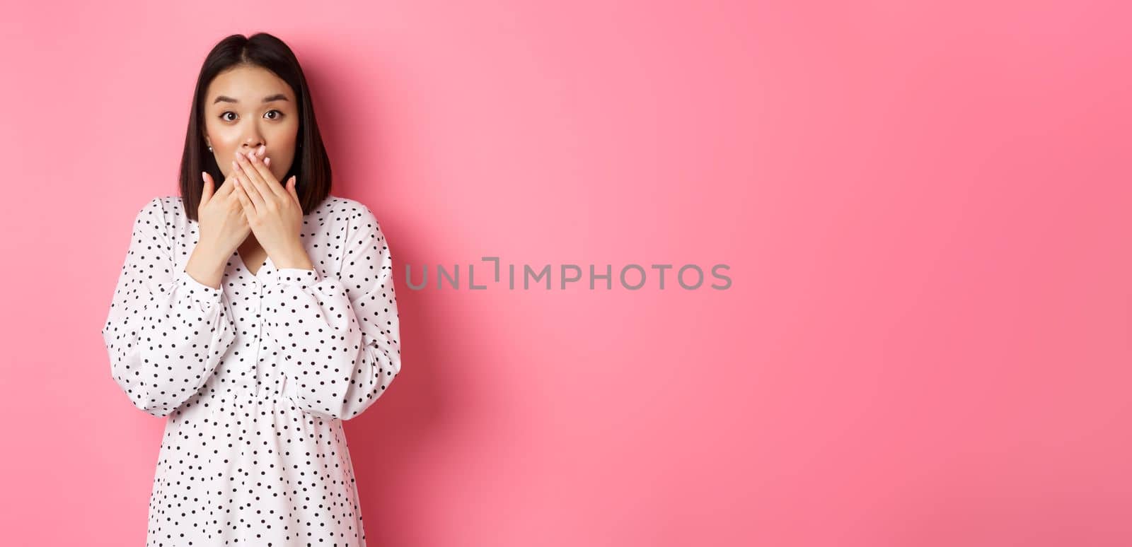 Image of surprised asian female model gasping, cover mouth and looking at camera, standing amazed against pink background by Benzoix