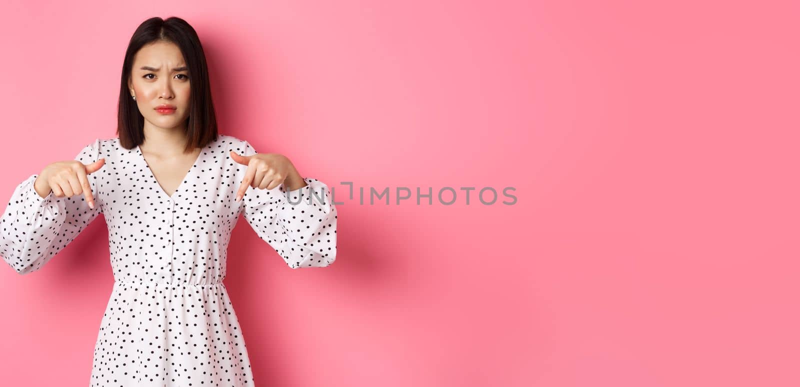 Disappointed asian female model pointing fingers down at promo offer, frowning and staring at camera upset, standing over pink background.