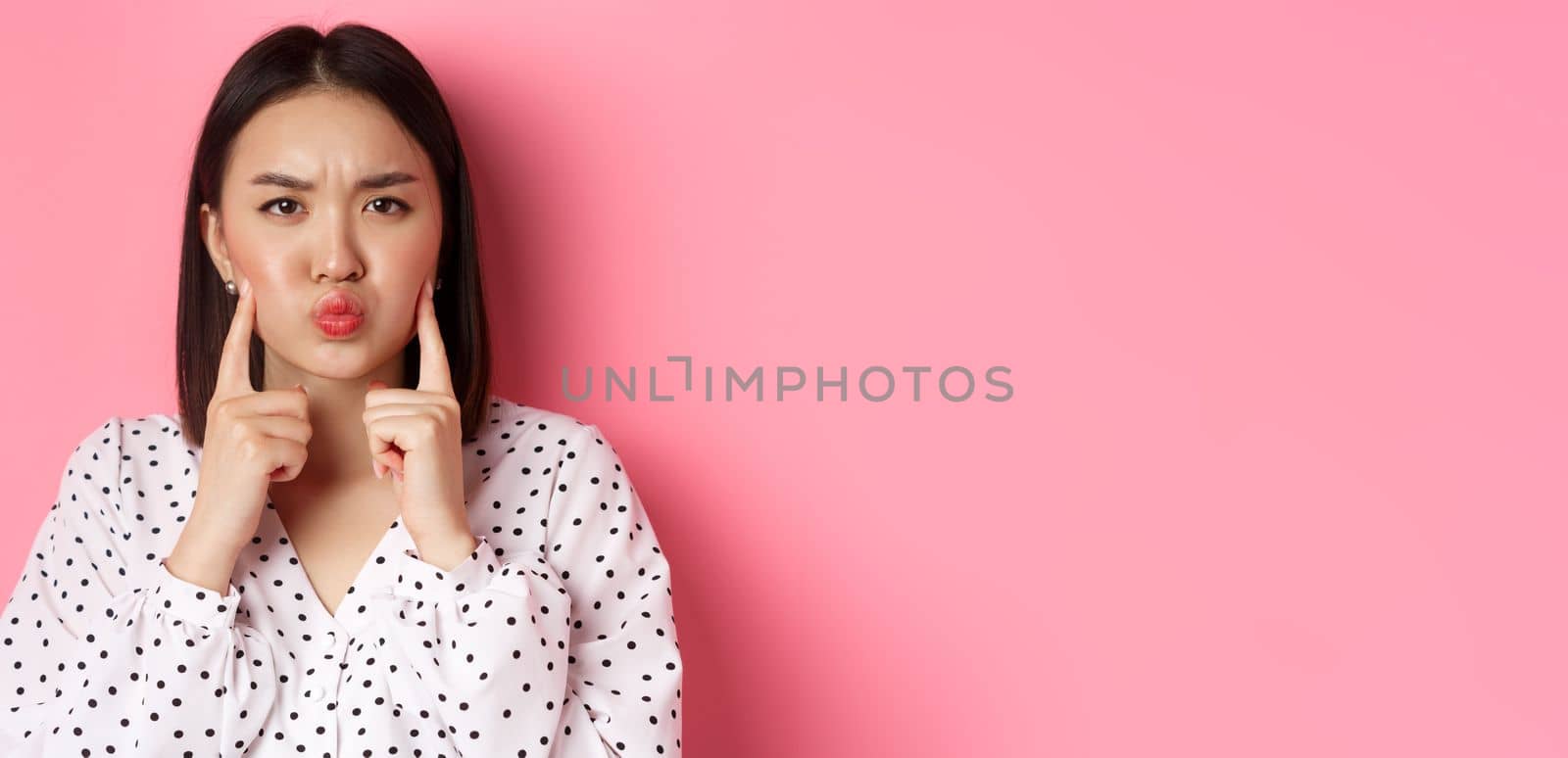 Beauty and lifestyle concept. Cute asian woman making grumpy face, poking cheeks and grimacing, standing over pink background.