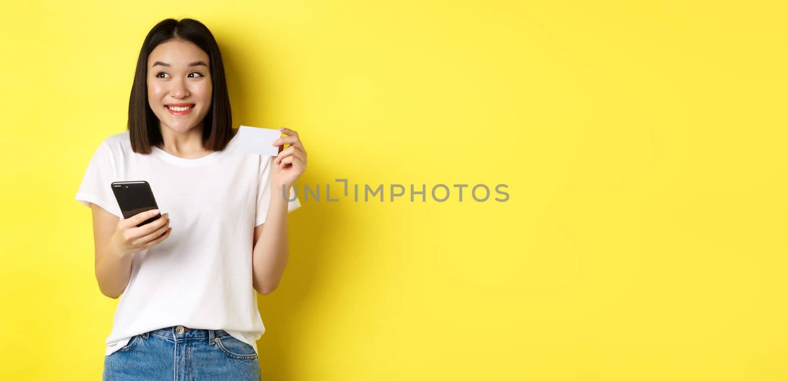 E-commerce and online shopping concept. Cheerful asian girl paying in internet, holding smartphone and plastic credit card, smiling and looking left, yellow background.