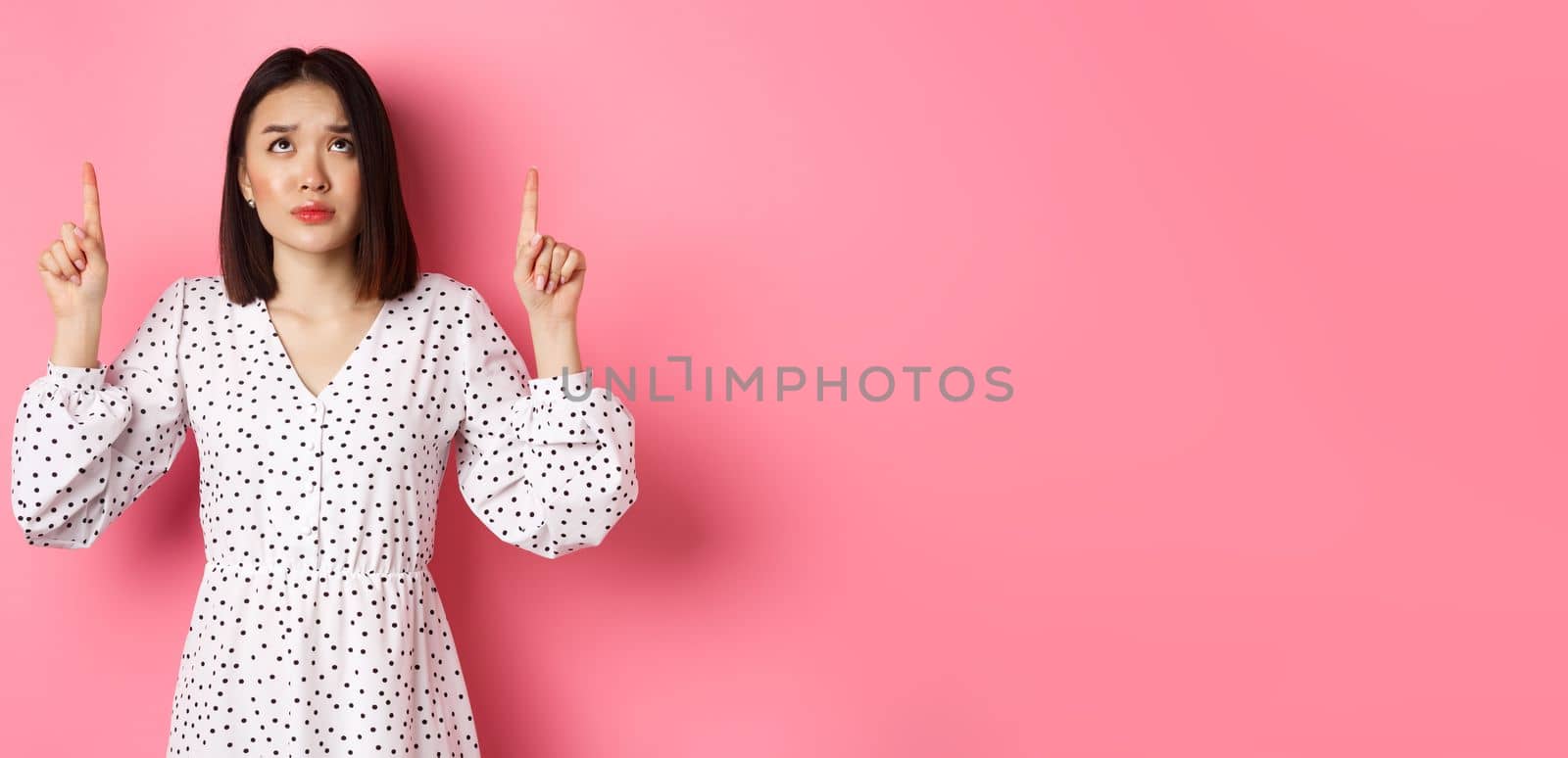 Worried and concerned asian girl in cute dress frowning, pointing and looking up with sad face, standing over pink background. Copy space