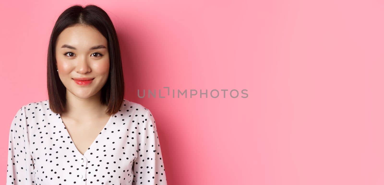 Close-up of cute asian female model looking at camera, blushing and smiling kawaii, standing over pink background.