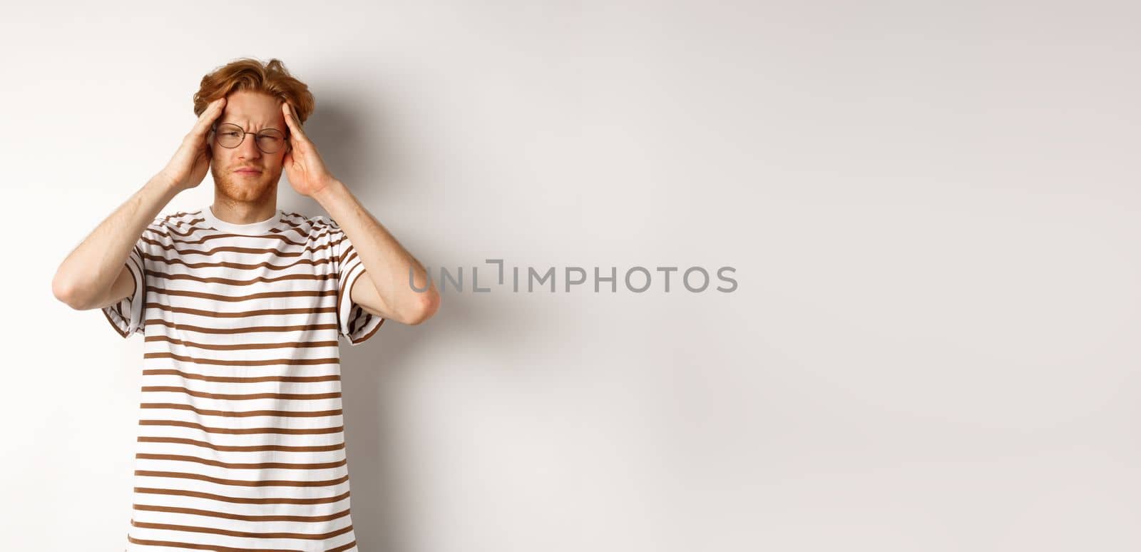 Image of young man with red hair and glasses touching head, frowning from painful migraine, having headache, standing over white background by Benzoix