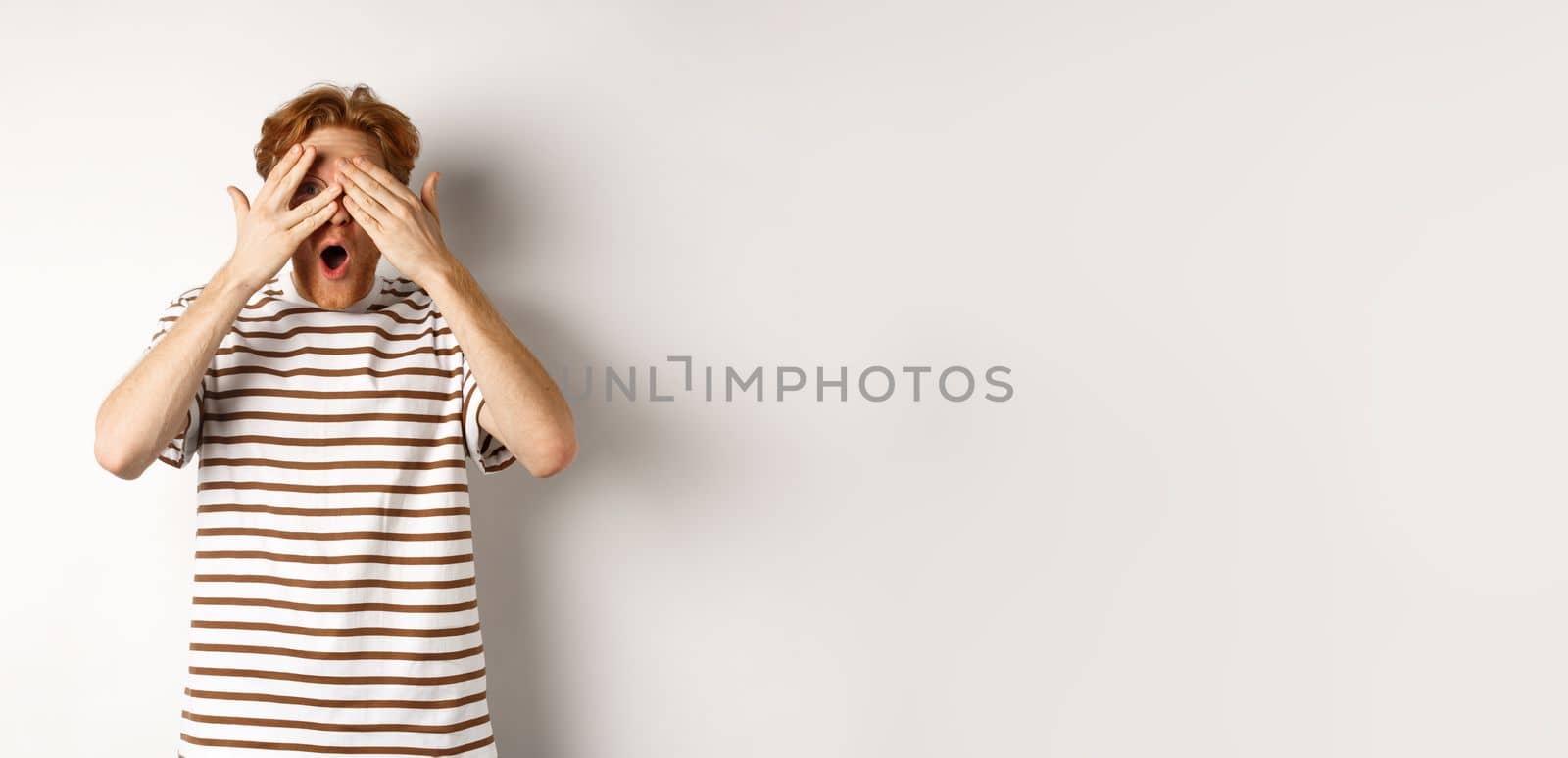 Shocked and embarrassed redhead guy covering eyes, peeking through fingers and gasping, standing over white background.