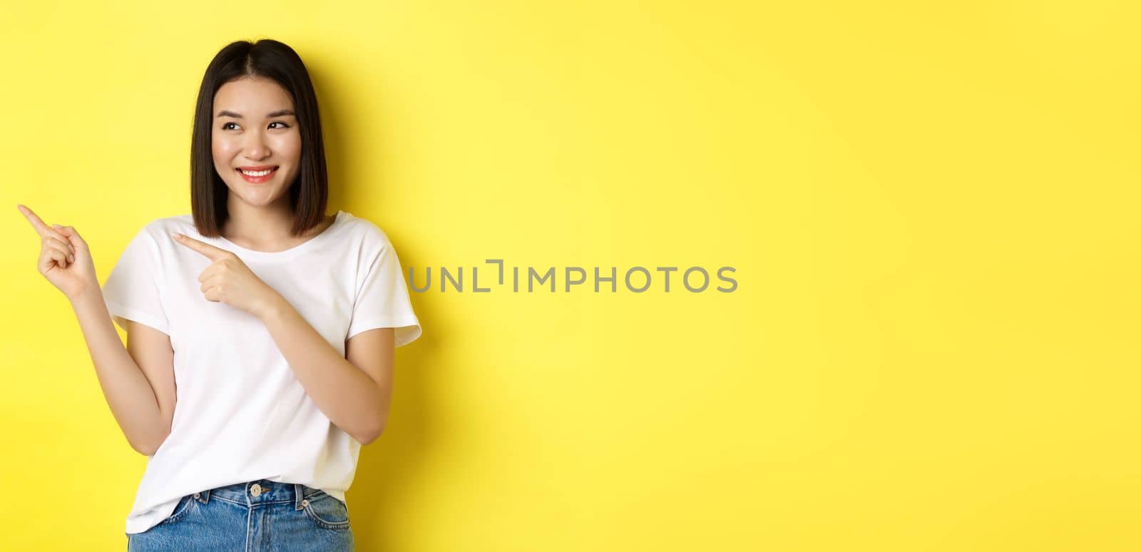Beauty and fashion concept. Beautiful asian woman in white t-shirt pointing fingers left, standing over yellow background.
