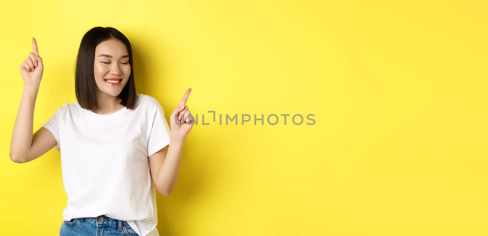 Happy asian woman dancing and having fun, posing in white t-shirt against yellow background.