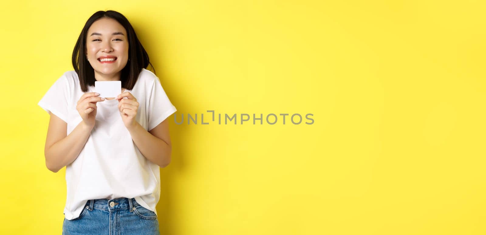 Young asian woman in casual white t-shirt going on shopping, showing plastic credit card and smiling excited, standing over yellow background by Benzoix