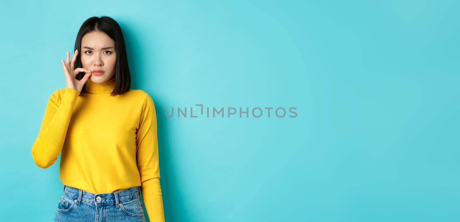 Serious asian girl showing mouth zip gesture, promise to keep shut and frowning, telling big secret, standing in yellow pullover over blue background.