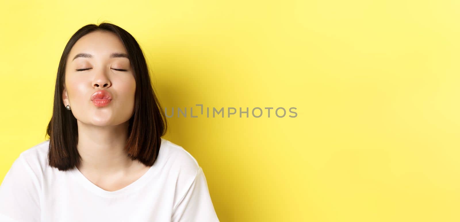 Valentines day concept. Close up of cute asian girl pucker lips and close eyes for kiss, standing romantic against yellow background by Benzoix