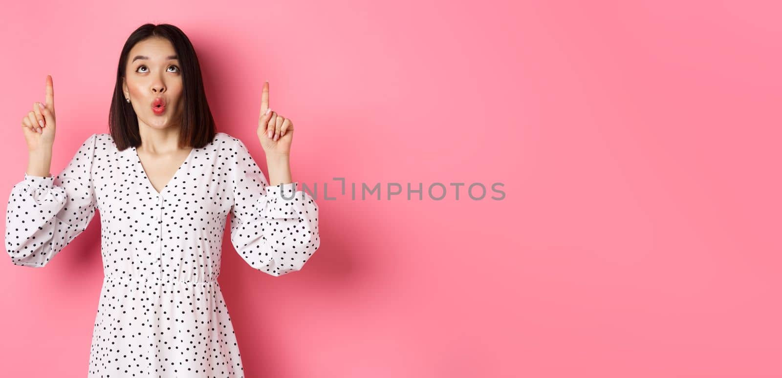 Cute korean girl in beautiful dress saying wow, looking and pointing fingers up, intrigued in promo offer, standing over pink background.
