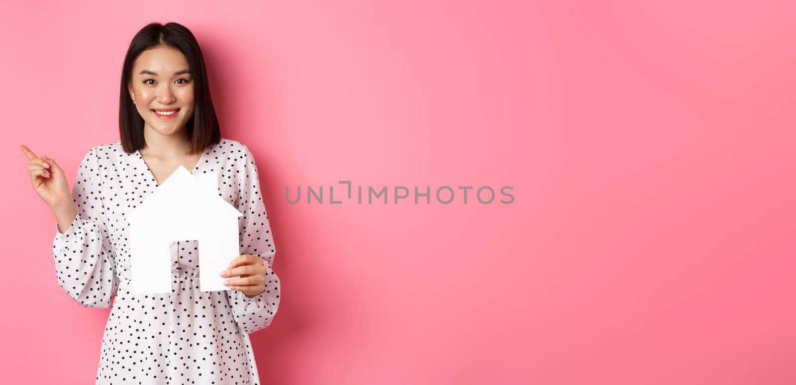Real estate. Excited asian woman showing paper house model, pointing left at copy space, standing over pink background.