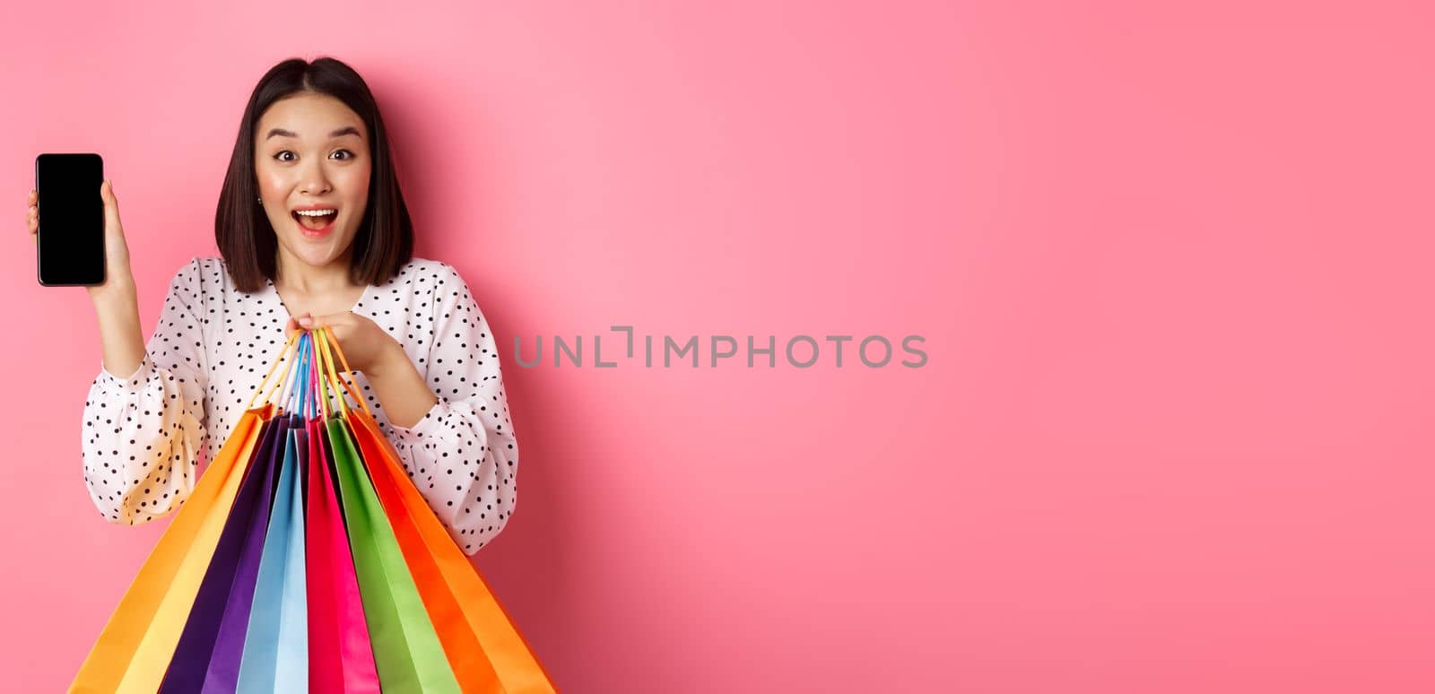 Attractive asian woman showing smartphone app and shopping bags, buying online via application, standing over pink background.