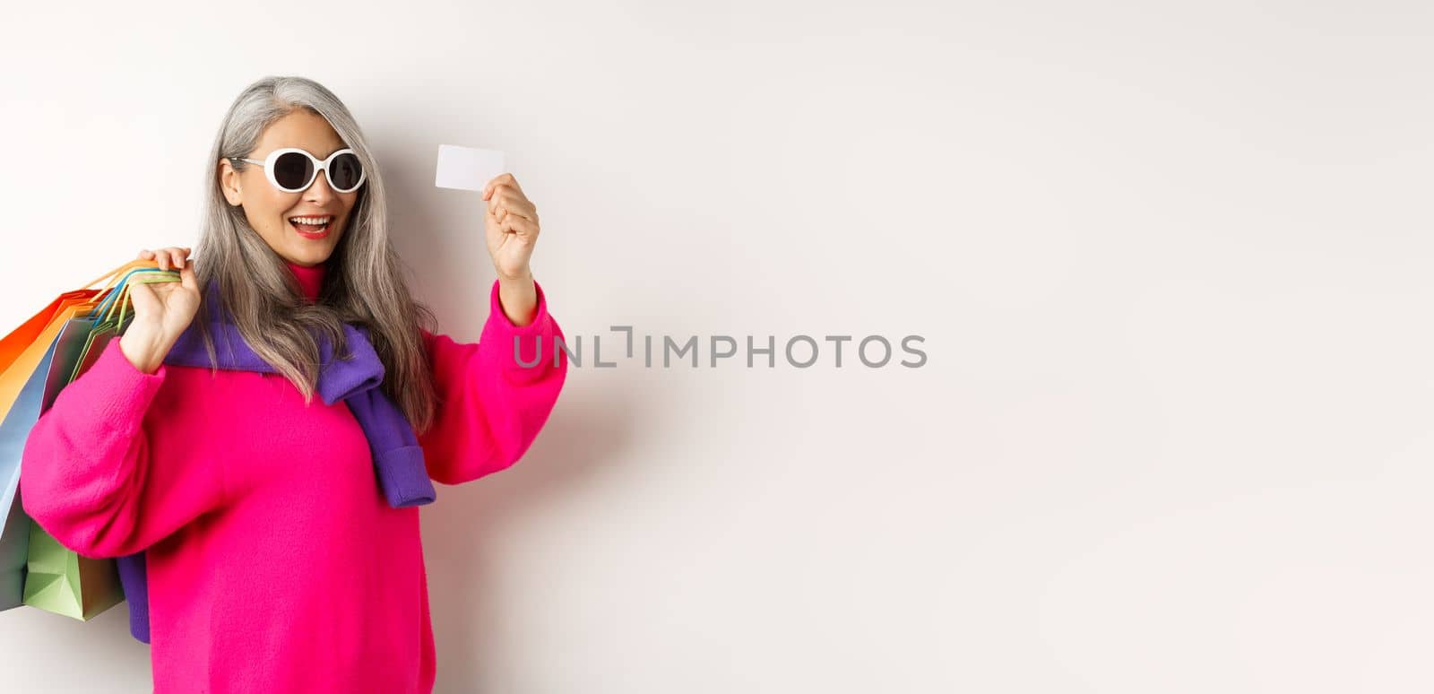 Fashionable asian old lady going shopping, holding paper bags and plastic credit card, wearing stylish sunglasses, standing over white background.