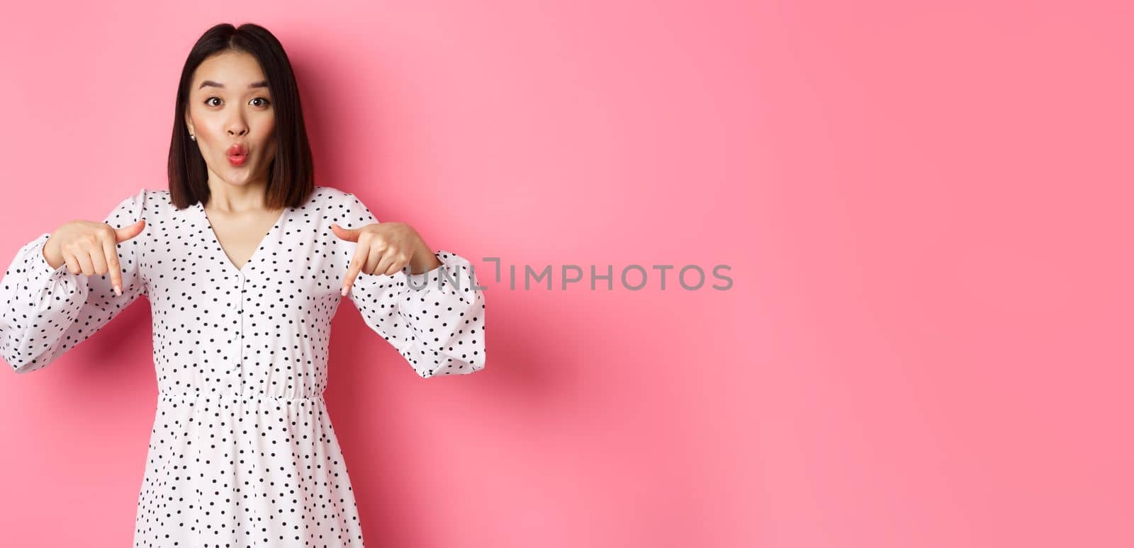 Impressed asian woman in dress pointing fingers down, look there gesture, check out discounts and sales, standing amazed over pink background by Benzoix