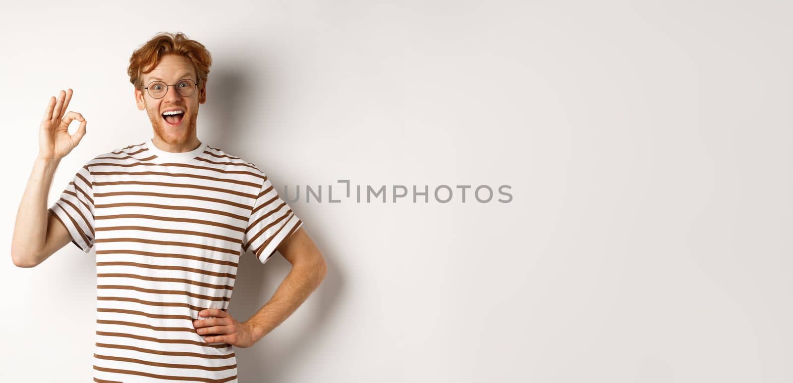 Excellent job. Happy redhead man praising you, showing OK sign and smiling amazed, approve and like something good, standing over white background by Benzoix