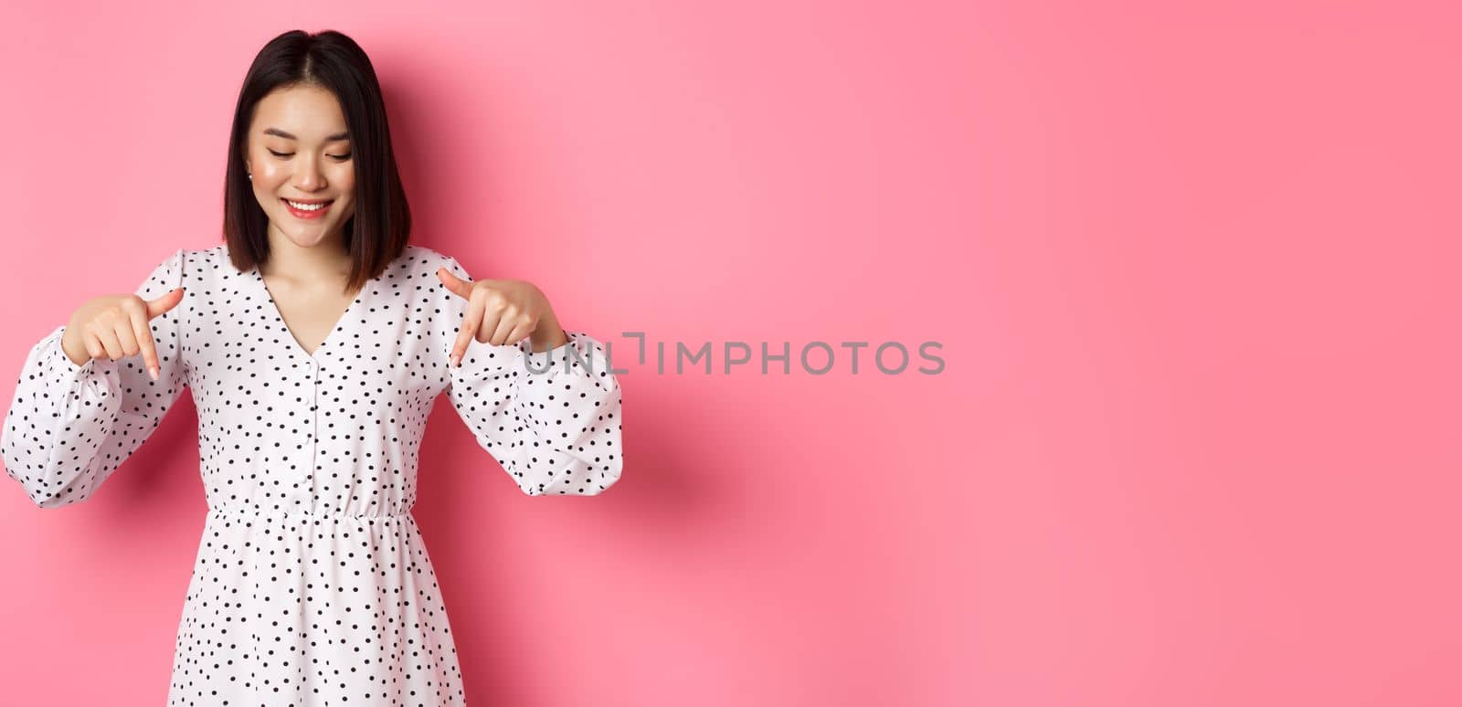 Cute romantic asian woman smiling, pointing fingers and looking down with pleased face, showing product discount, standing over pink background.