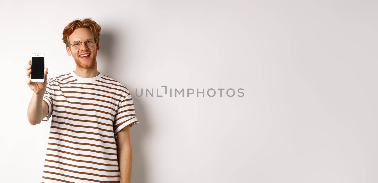 Technology and e-commerce concept. Happy young redhead man in glasses showing blank smartphone screen, smiling satisfied, standing over white background.