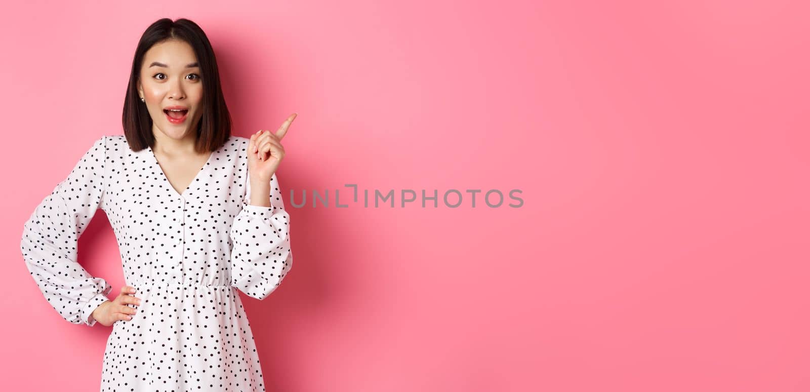 Excited pretty asian girl pointing at upper right corner, gasping amazed and looking at camera, discuss store promo offer, standing over pink background.