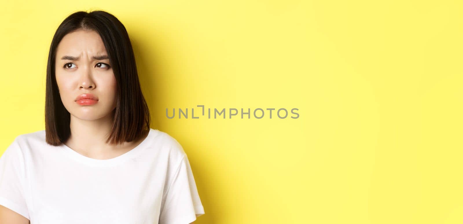 Sad and jealous asian girl sulking, frowning and looking left with upset face, standing over yellow background.