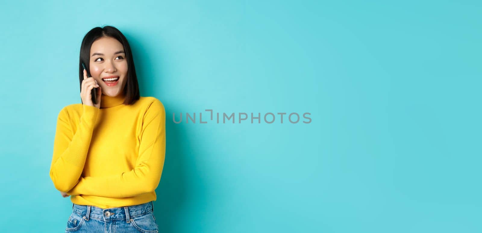 Stylish korean woman calling on phone, talking on smartphone and looking happy at upper right corner, smiling carefree, standing in relaxed pose over blue background by Benzoix