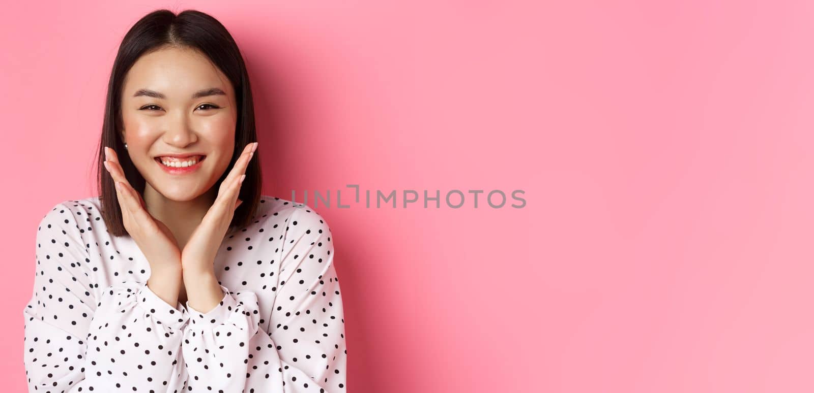 Beauty and skin care concept. Close-up of cute asian woman showing clean perfect face and smiling, looking happy at camera, standing over pink background.