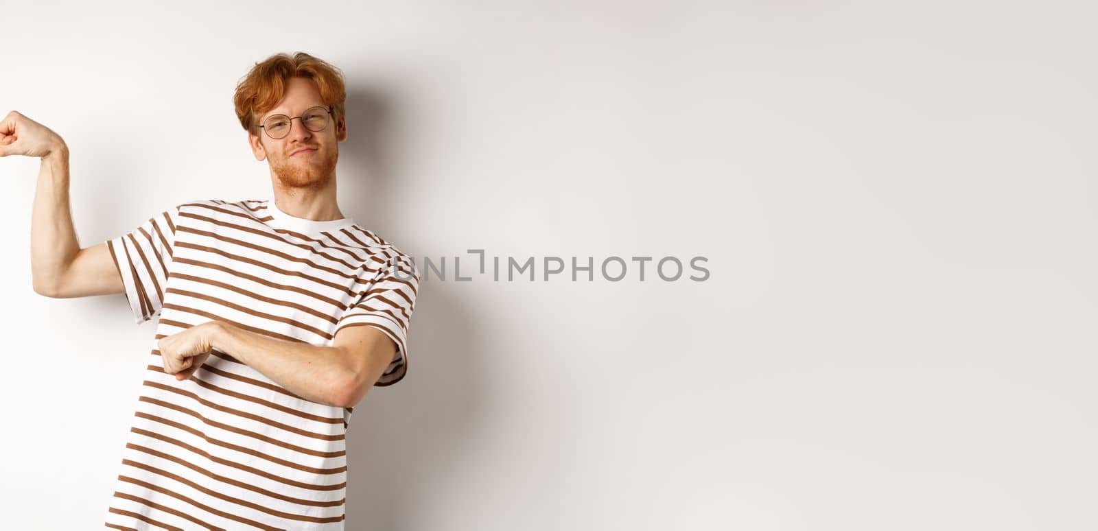 Image of confident and strong redhead man flexing biceps, showing muscles after gym, standing over white background.