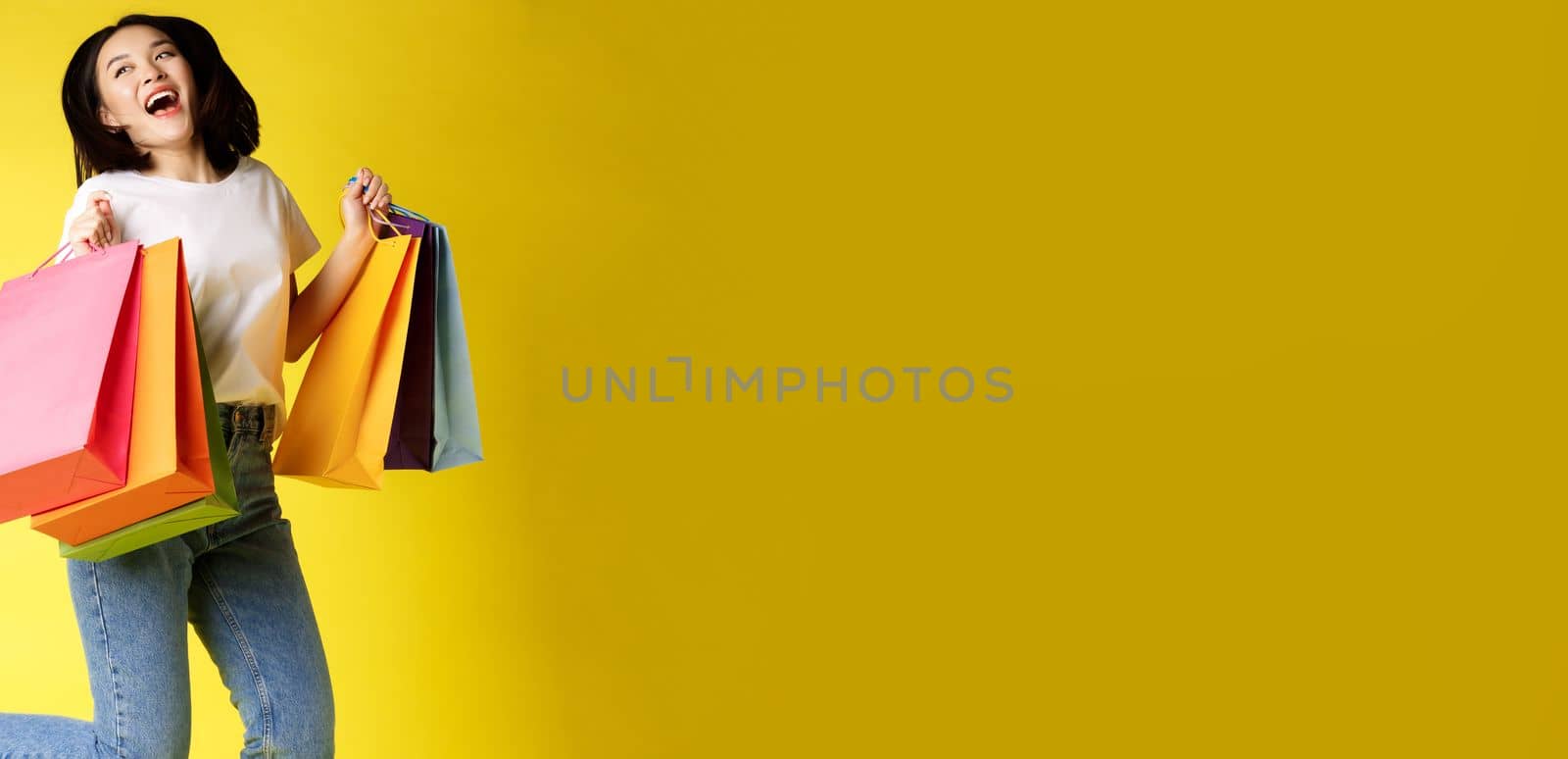 Full size shot of beautiful asian woman jumping with shopping bags and smiling happy, yellow background.