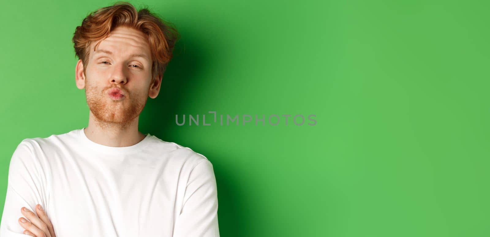 Emotions and fashion concept. Silly redhead guy with beard pucker lips and leaning for kiss, standing over green background.