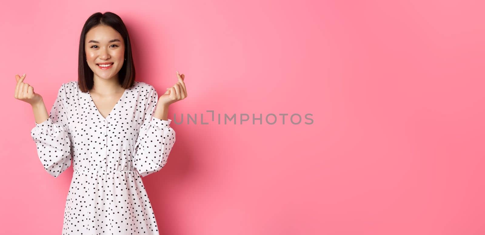 Lovely asian woman in dress showing korean heart signs and smiling, standing on romantic pink background by Benzoix