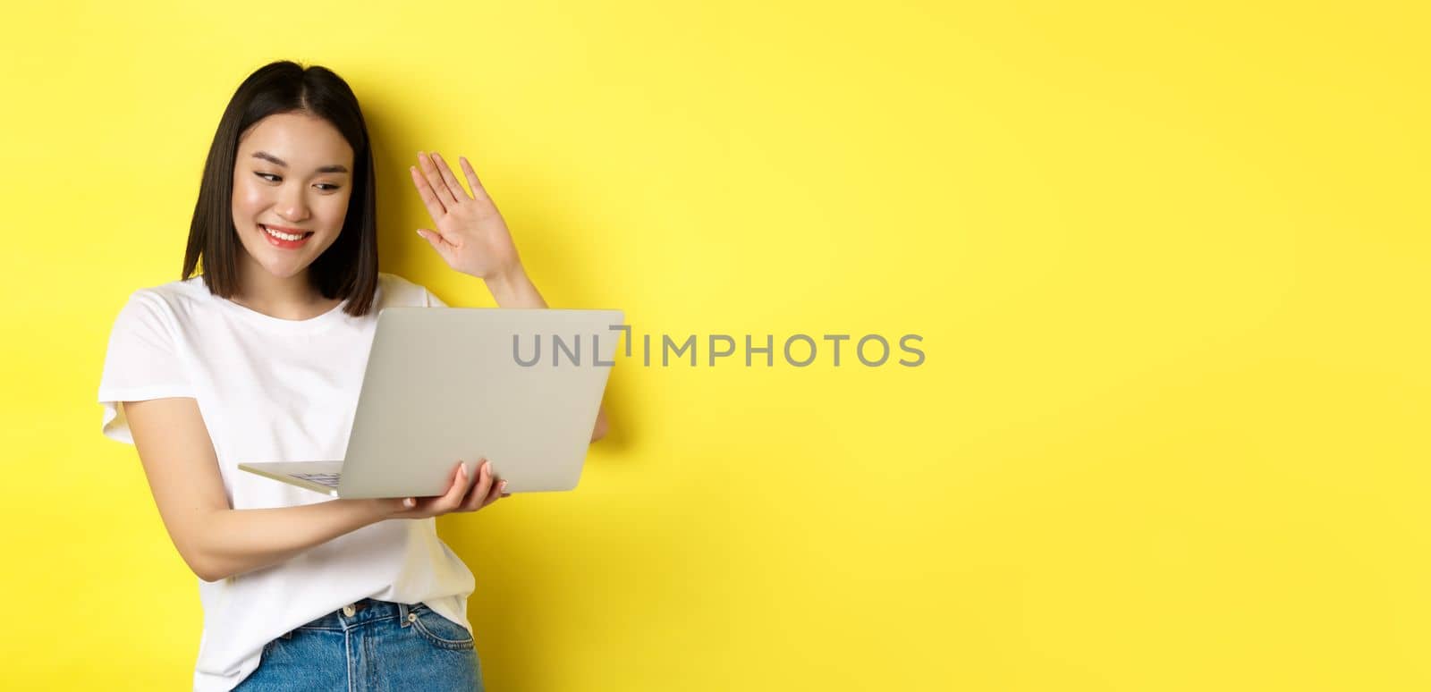 Happy asian woman video chat on laptop, waiving hand at computer camera and saying hello, standing over yellow background.