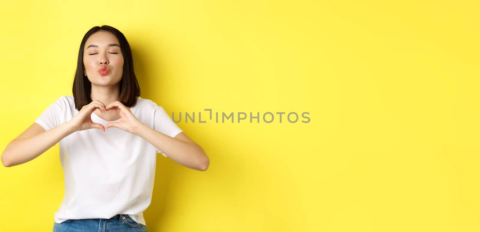 Beautiful asian woman showing I love you heart gesture, smiling at camera, standing against yellow background. Concept of valentines day and romance.