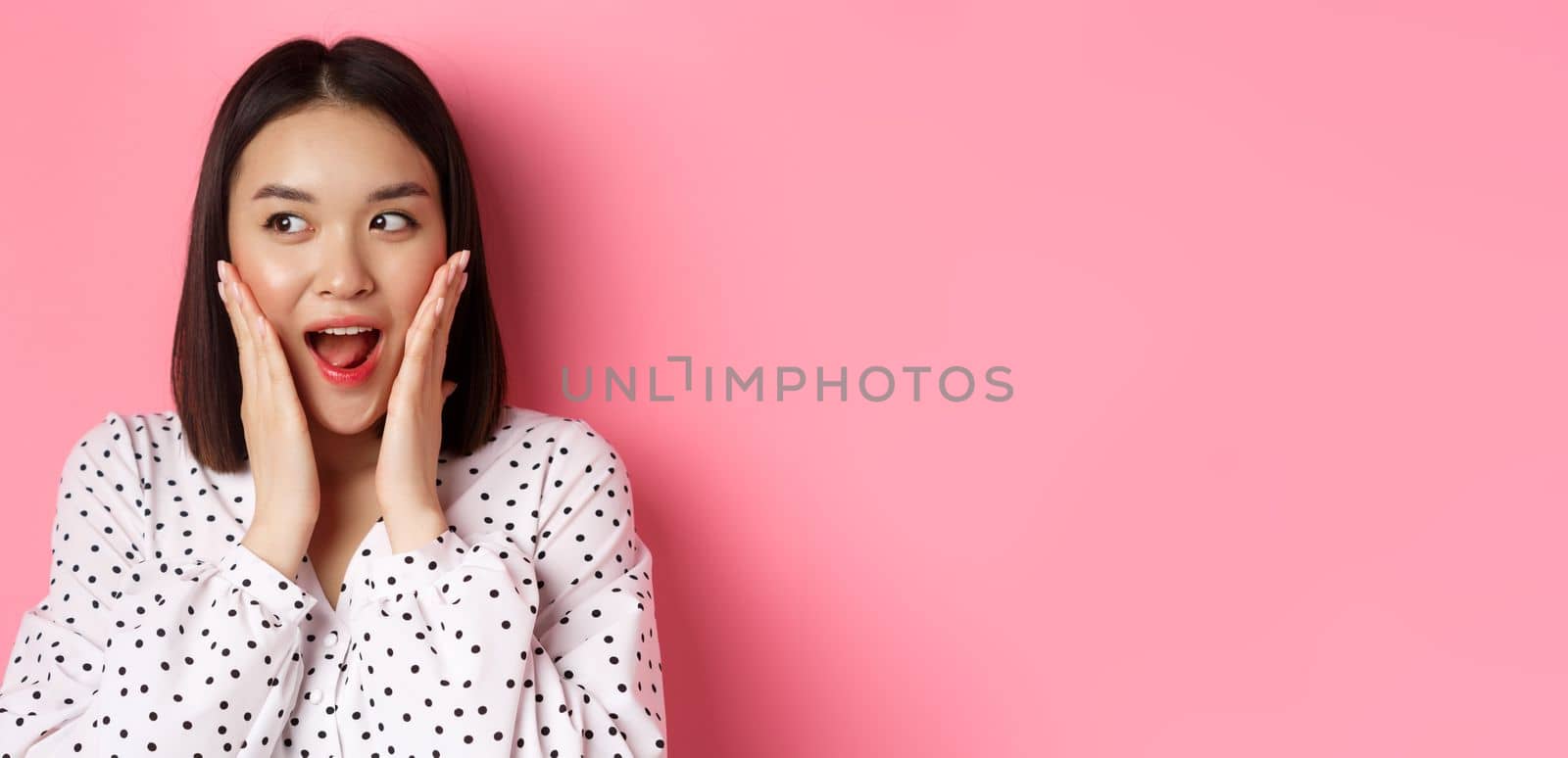 Close-up of beautiful asian woman looking surprised and excited, hear amazing news, looking left and rejoicing, standing against pink background.