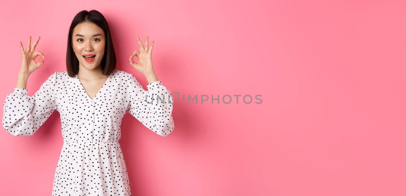 Pretty young asian woman in dress showing okay sign, praising and showing approval, looking satisfied, standing against pink background by Benzoix