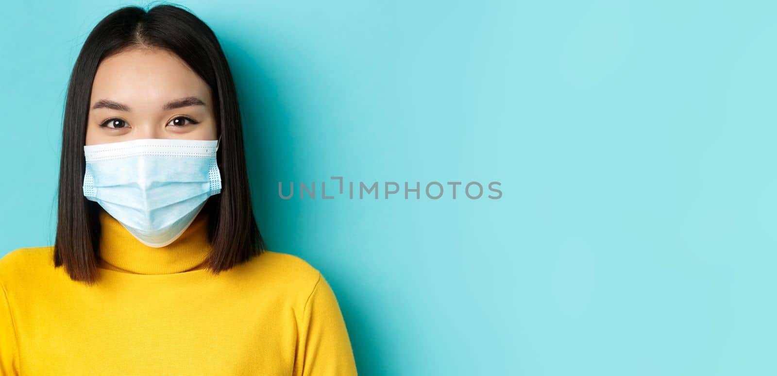 Covid-19, social distancing and pandemic concept. Close up of young asian woman with short dark hair, wearing medical mask and smiling with eyes, looking hopeful at camera by Benzoix