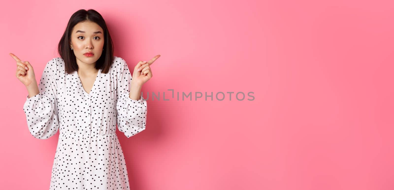 Confused asian woman hesitating, shrugging and pointing hands two sides, staring indecisive and need help with choice, standing over pink background.