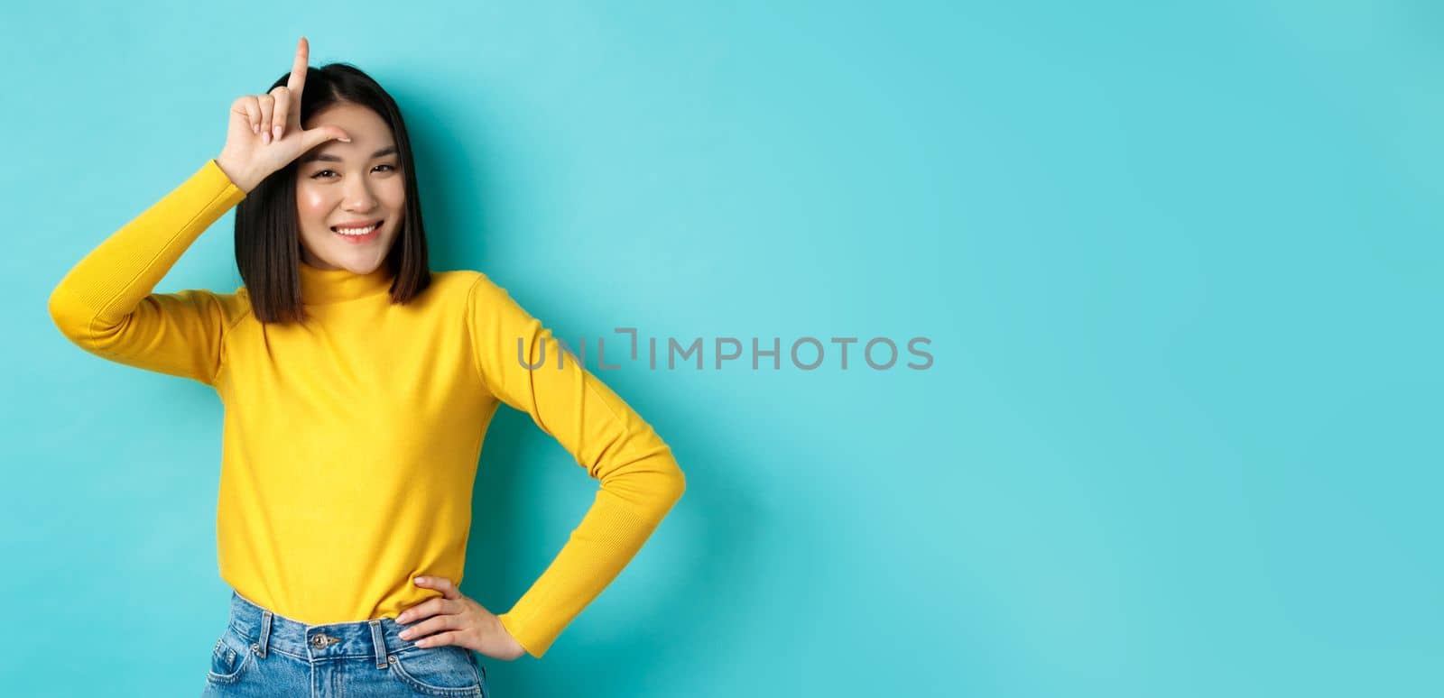 Sassy asian girl mocking lost team, showing loser sign on forehead and smiling pleased, being a winner, standing over blue background.