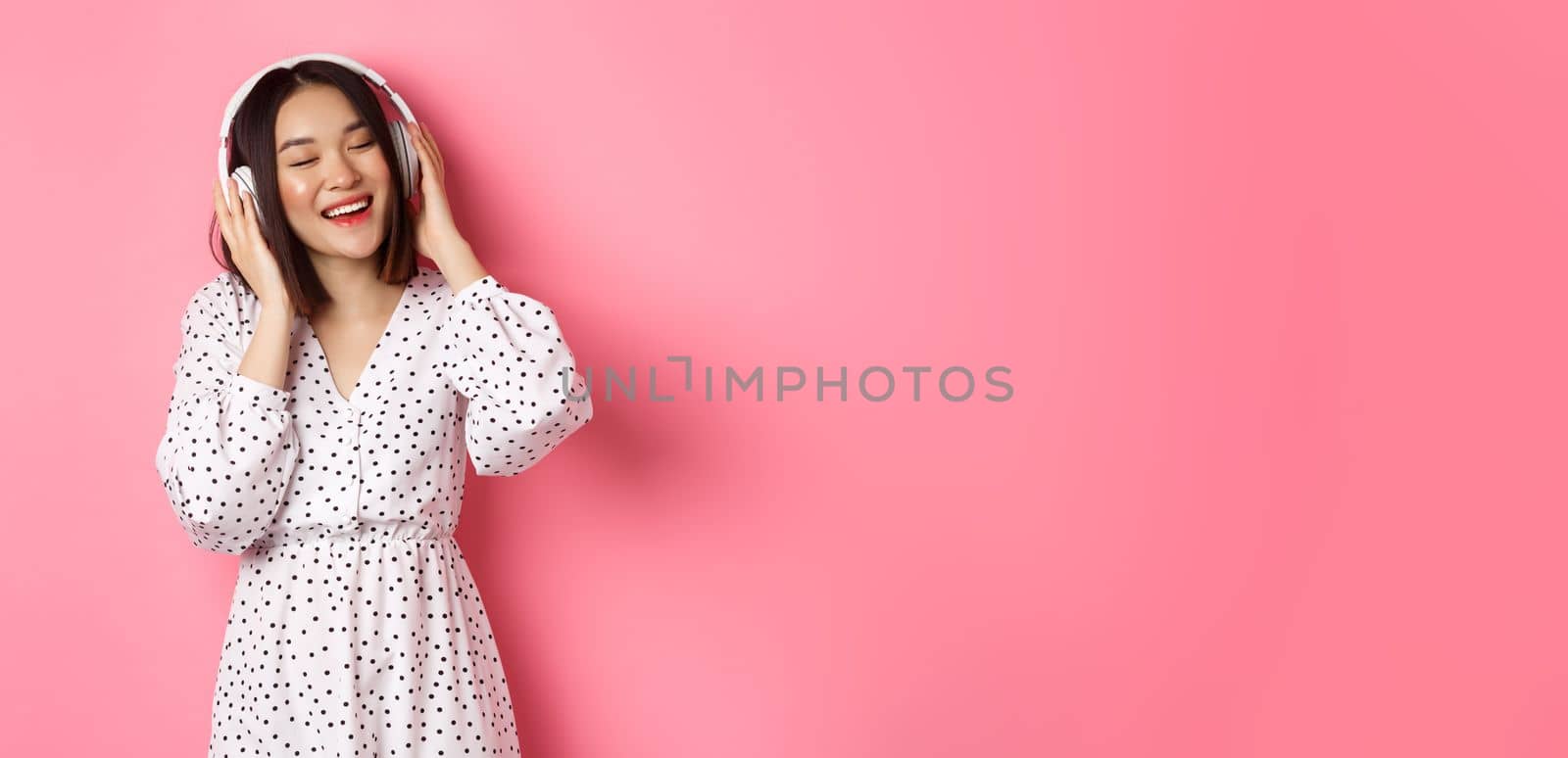 Romantic asian woman smiling happy, listening music in headphones and dancing, standing in trendy dress over pink background by Benzoix