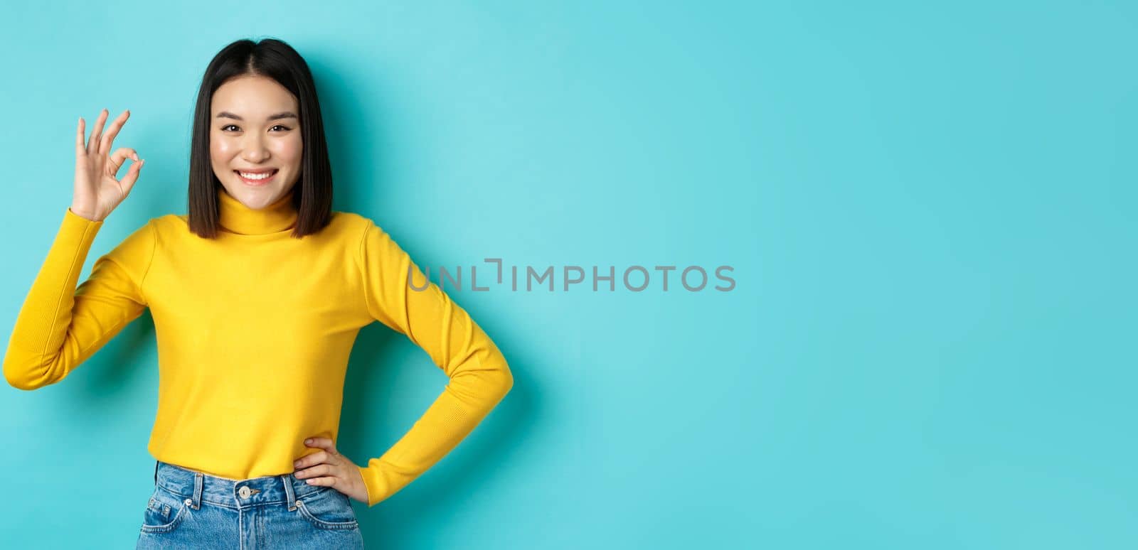 Happy beautiful asian woman smiling, showing okay gesture in approval, saying yes, standing pleased over blue background by Benzoix