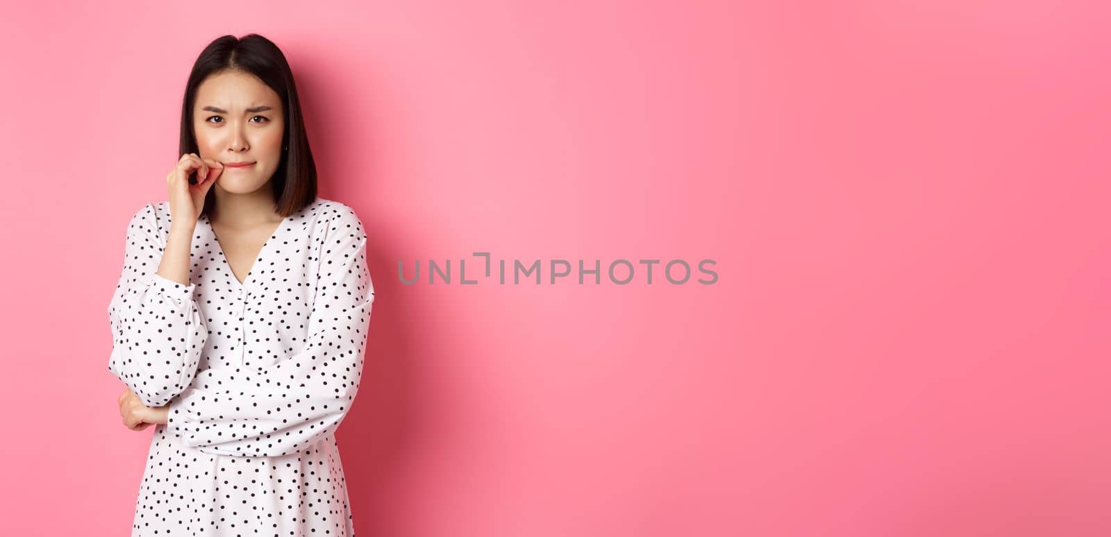Brunette asian woman in dress looking displeased, frowning and zipping mouth, seal lips with promise, standing over pink background.